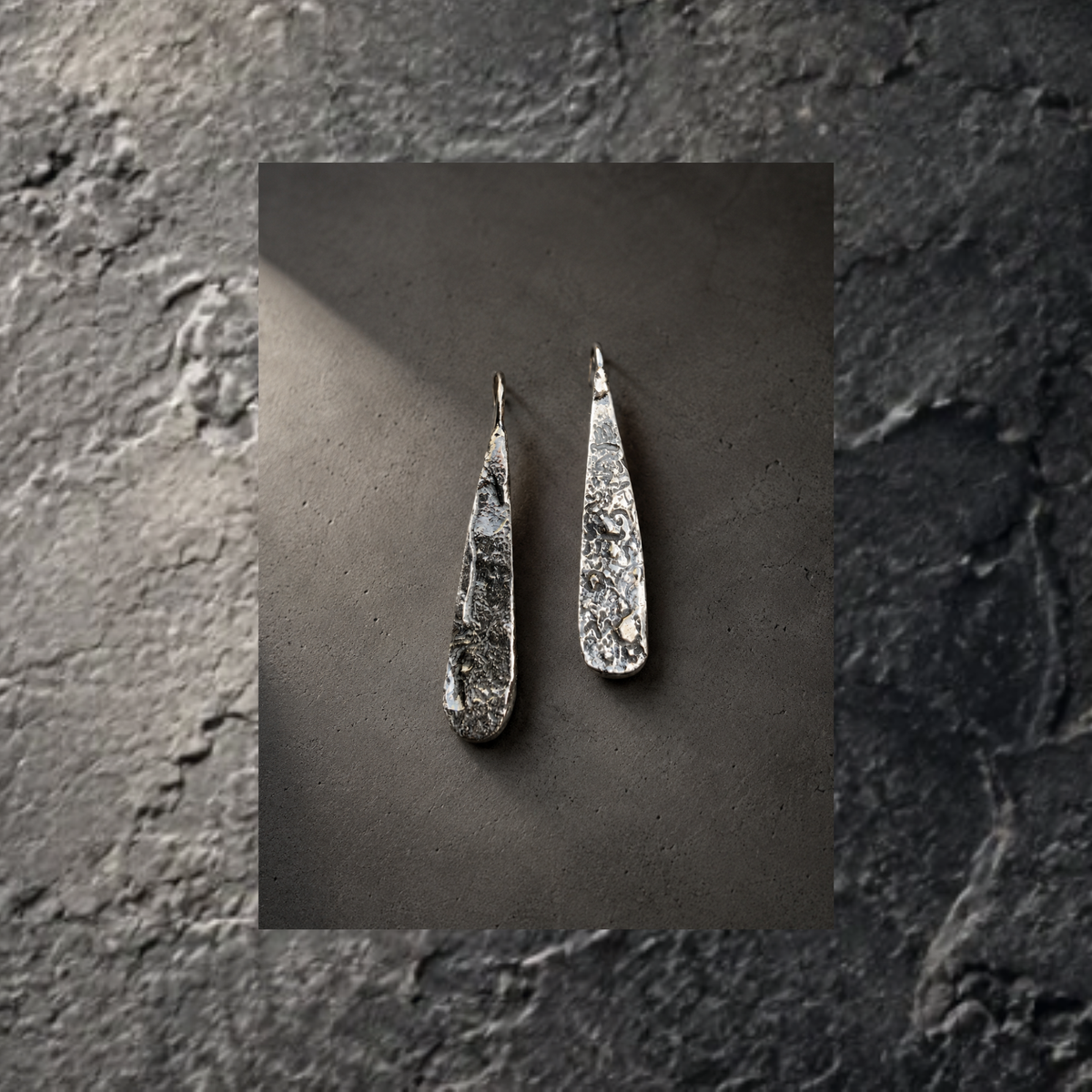 A pair of textured silver teardrop dangle earrings displayed on a dark gray textured background, presented as a centered square print against a dark stone-textured backdrop.