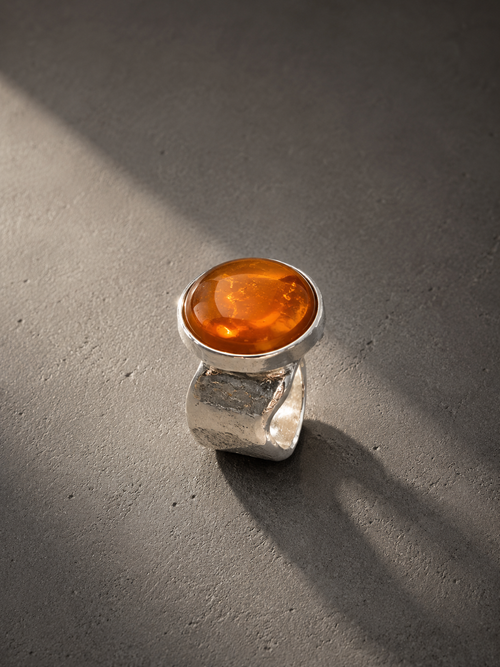 A silver ring with a large, polished oval amber cabochon gemstone, photographed on a textured gray concrete surface under dramatic, directional sunlight.