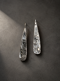 A pair of hammered silver dangle earrings with a textured organic surface, displayed on a dark gray textured background.