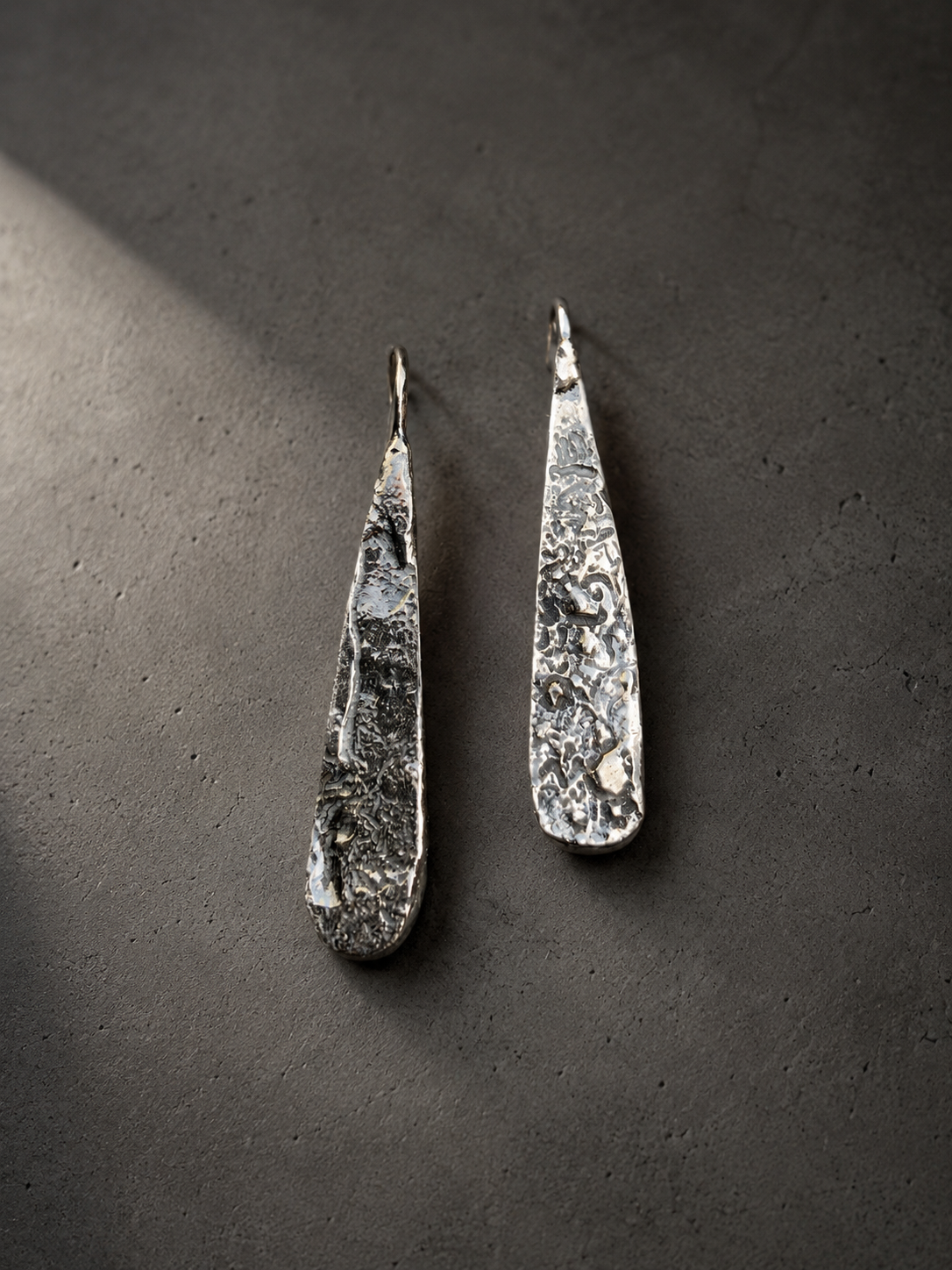 A pair of hammered silver dangle earrings with a textured organic surface, displayed on a dark gray textured background.