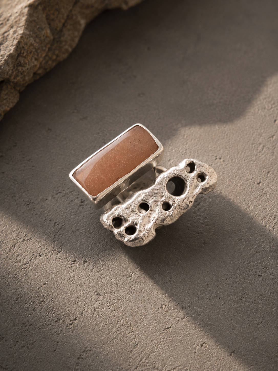 A silver ring featuring a rectangular warm brown stone set on top of a chunky, textured silver band with organic holes, photographed on a textured gray surface with a rock in the corner.