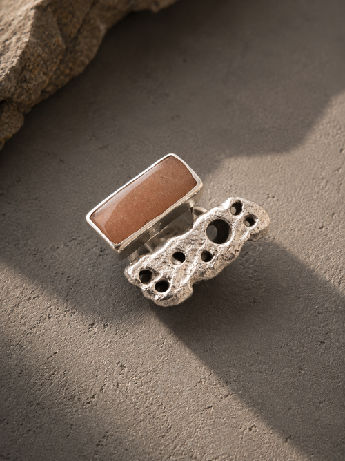 A silver ring featuring a rectangular warm brown stone set on top of a chunky, textured silver band with organic holes, photographed on a textured gray surface with a rock in the corner.