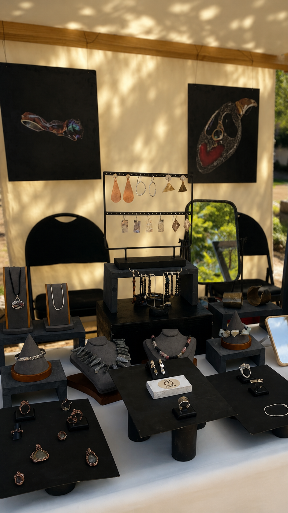 A jewelry display booth with multiple black stands, necklaces, rings, earrings, and bracelets arranged on a table under a canopy. Two black wall art pieces hang above the display, and a mirror reflects the setup.