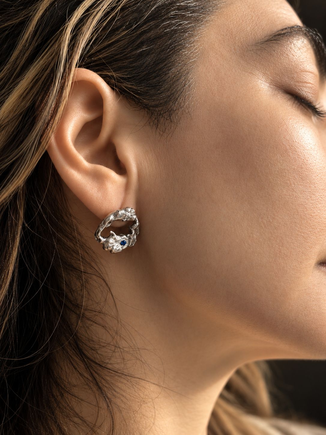 A close-up side profile of a woman wearing a silver-toned textured hoop earring with a small dark blue gemstone accent.