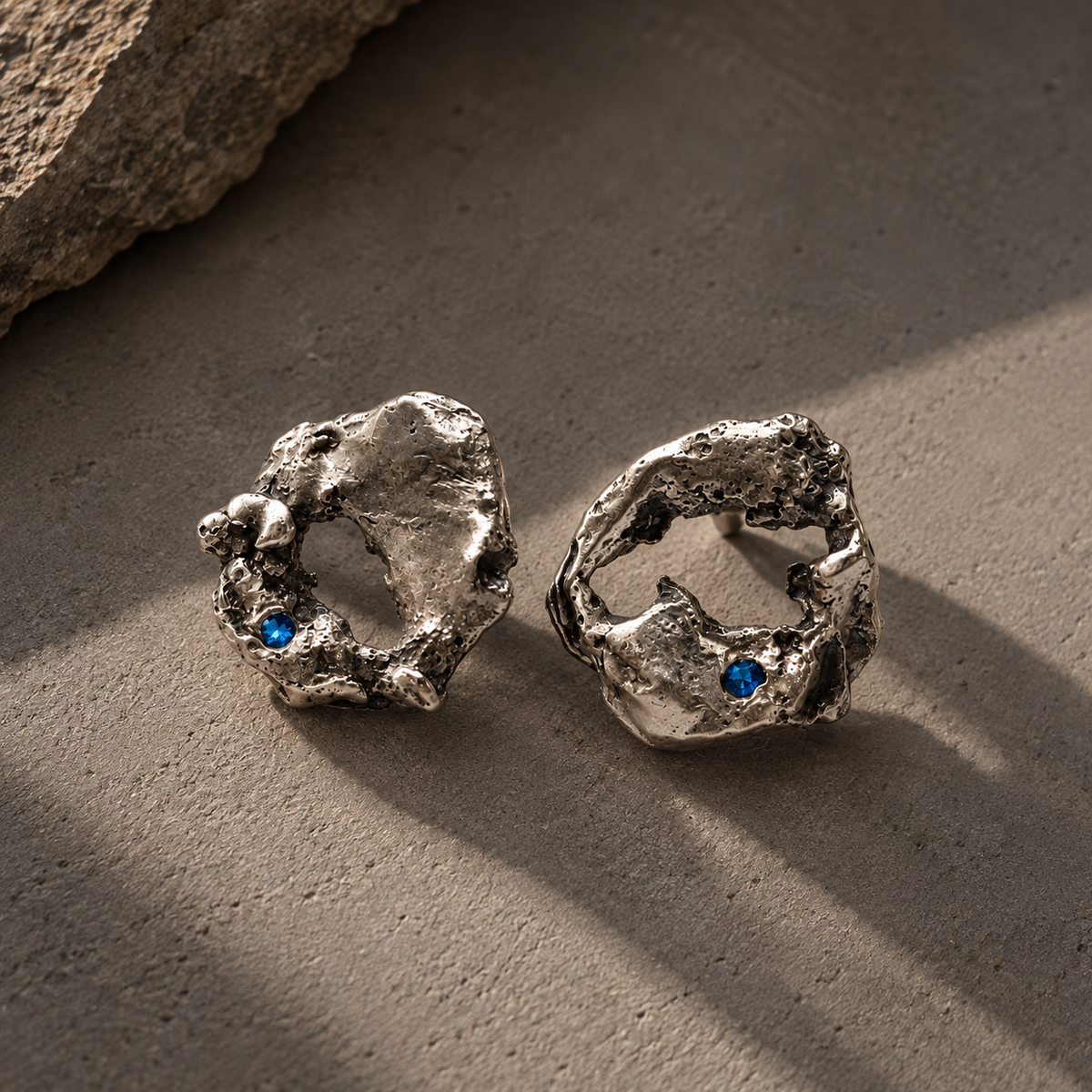 A pair of textured silver earrings with organic, rough organic shapes and small blue gemstone accents, displayed on a neutral concrete surface with natural shadows.